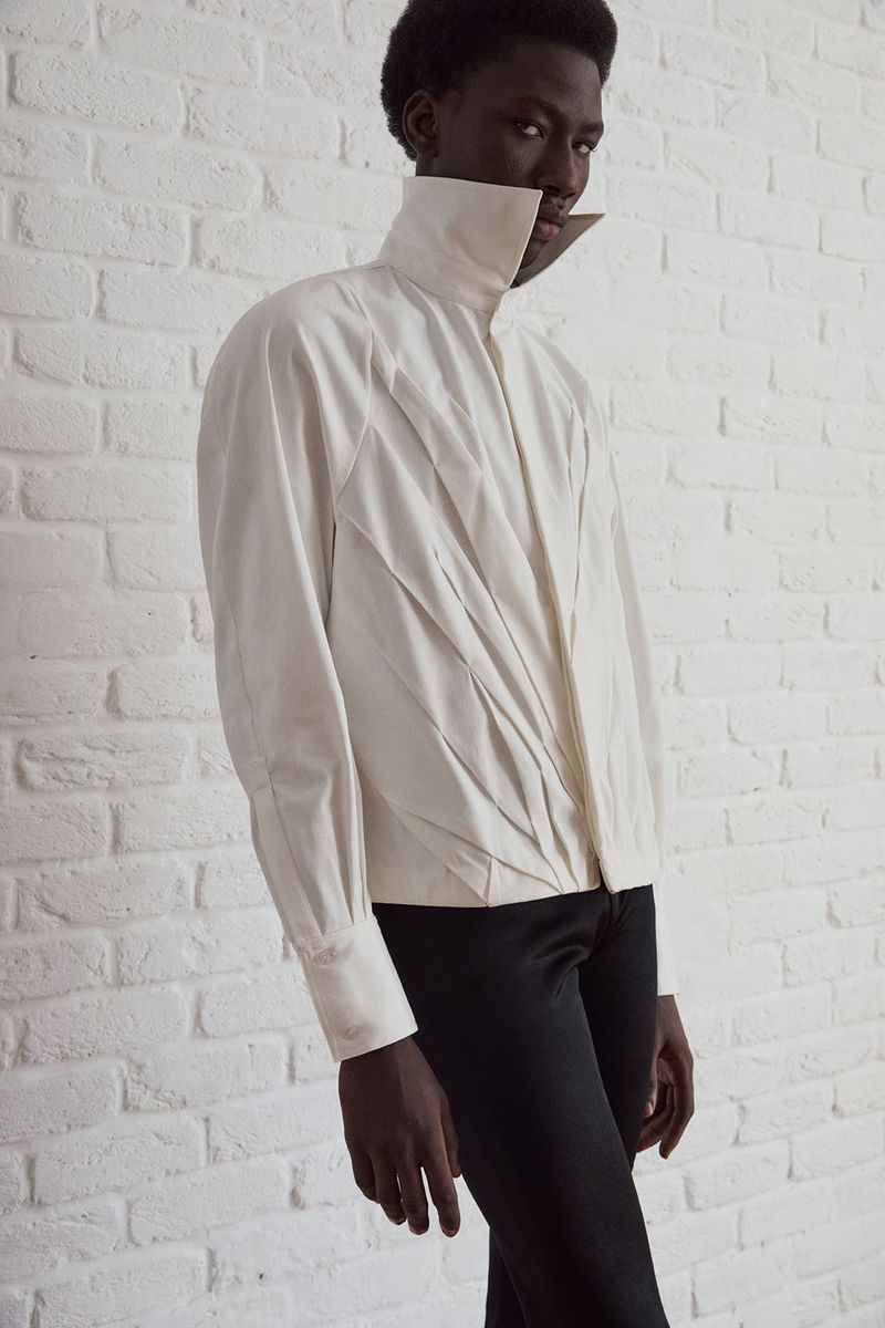 Dark-skinned model wearing an off-white, high-collar shirt and black pants stands in a studio setting with a white brick wall backdrop.