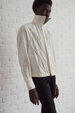 Dark-skinned model wearing an off-white, high-collar shirt and black pants stands in a studio setting with a white brick wall backdrop.