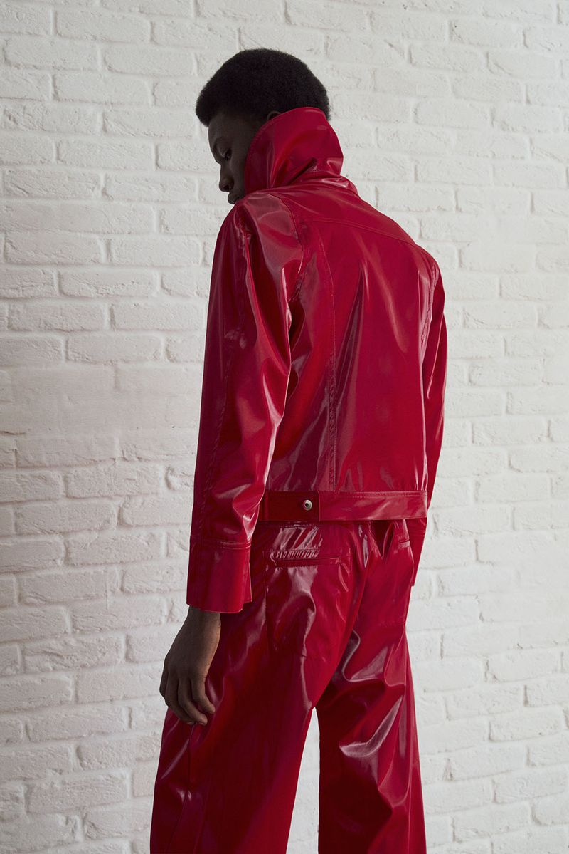 Model in shiny red jacket and pants against white brick wall. Fashion editorial portrait. Bright studio lighting. Minimalist urban aesthetic.