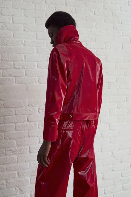 Model in shiny red jacket and pants against white brick wall. Fashion editorial portrait. Bright studio lighting. Minimalist urban aesthetic.
