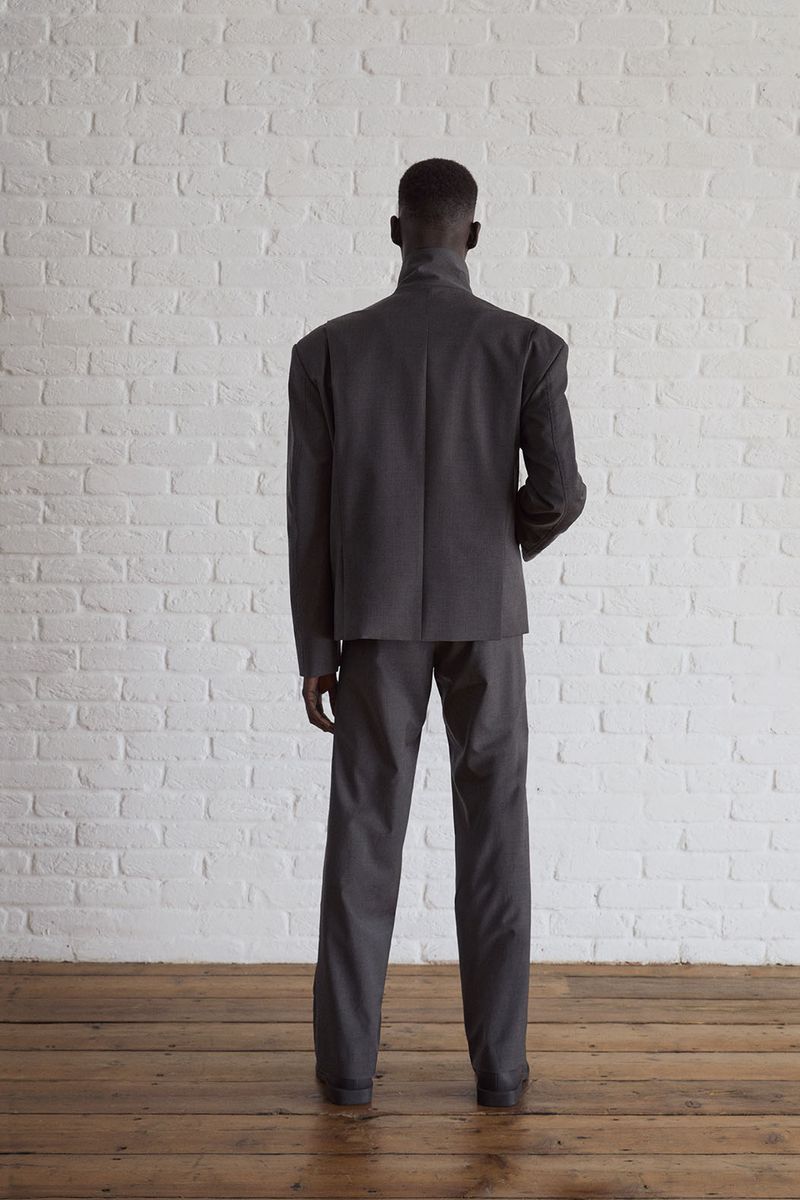 A slender adult male stands indoors with his back to the camera, wearing a dark grey suit against a white brick wall backdrop.