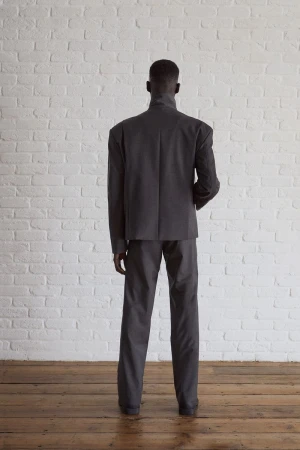 A slender adult male stands indoors with his back to the camera, wearing a dark grey suit against a white brick wall backdrop.