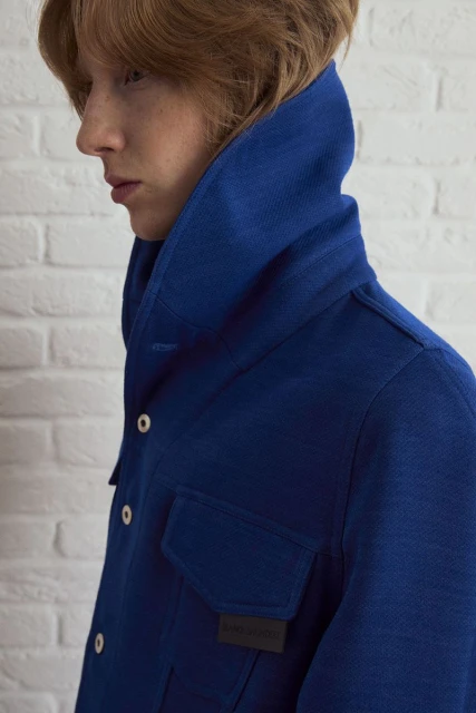 A person with red hair wears a royal blue jacket in a studio headshot against a white brick wall, showcasing a fashion-forward aesthetic.