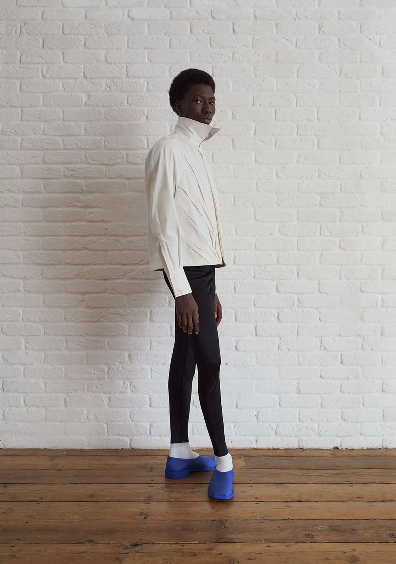 Model standing in profile wearing a cream high-collar jacket, black slim trousers, and bright blue shoes against a white brick wall.