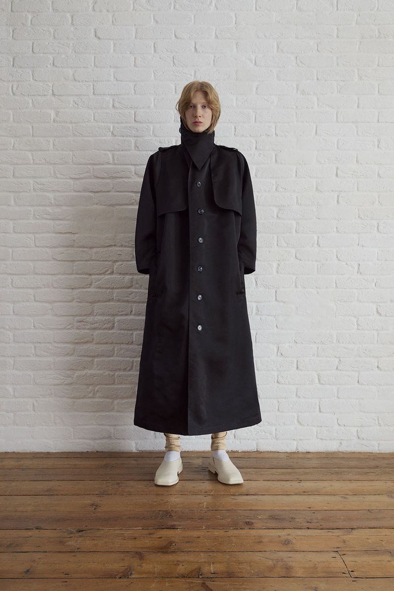 Full-length studio shot of a fair-skinned adult woman in a long black coat, high collar, beige leggings, and white shoes against white brick wall.