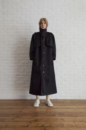 Full-length studio shot of a fair-skinned adult woman in a long black coat, high collar, beige leggings, and white shoes against white brick wall.