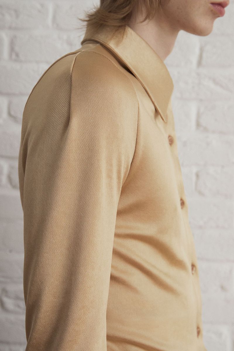 Close-up studio shot of an adult wearing a beige button-down shirt with a collar, against a textured white wall background.