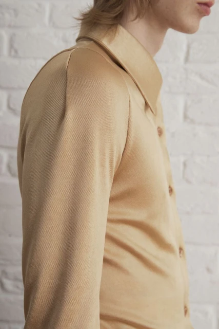 Close-up studio shot of an adult wearing a beige button-down shirt with a collar, against a textured white wall background.