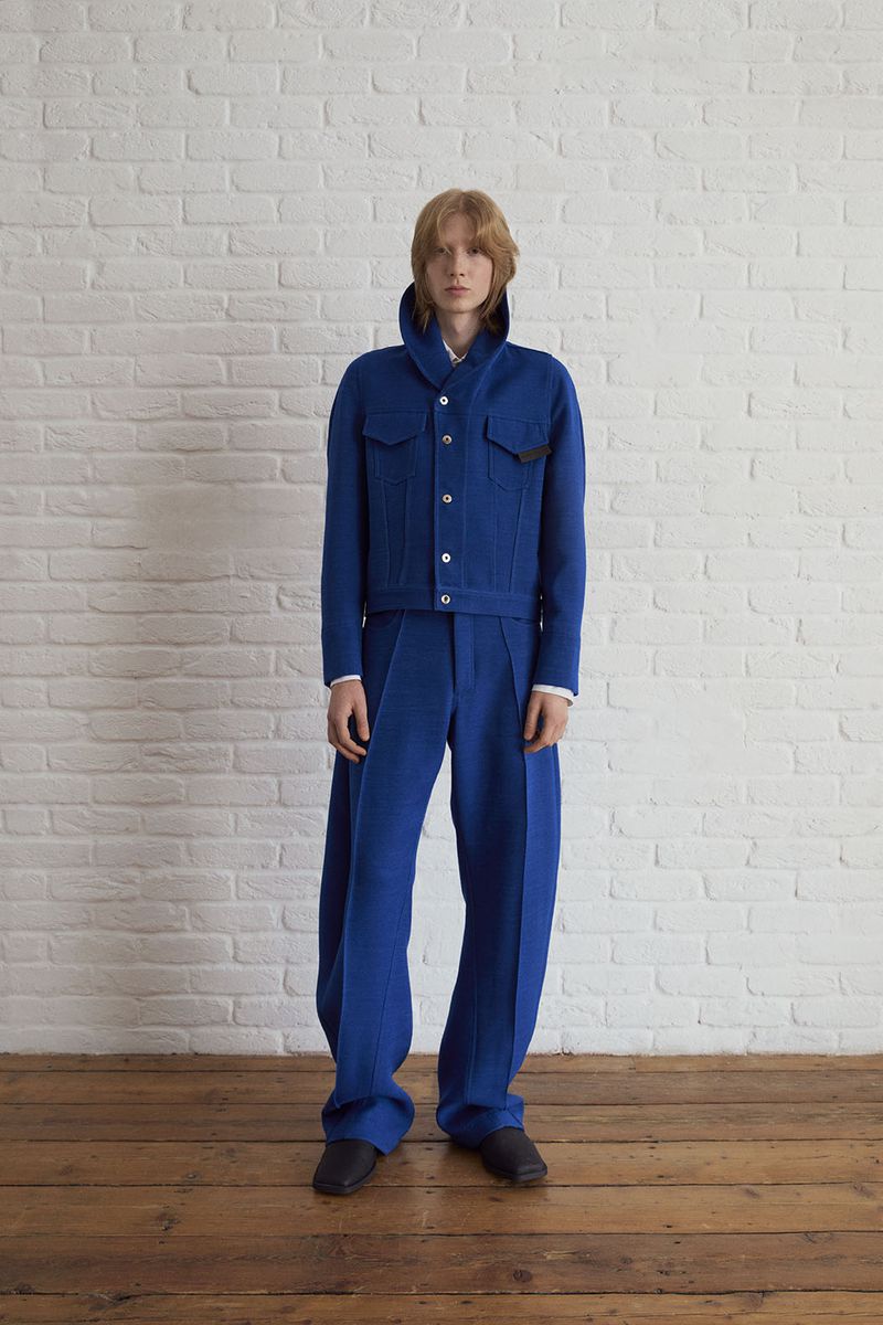 Young adult male fashion model stands in a photo studio wearing a matching blue wool jacket and wide-leg pants in a full shot.