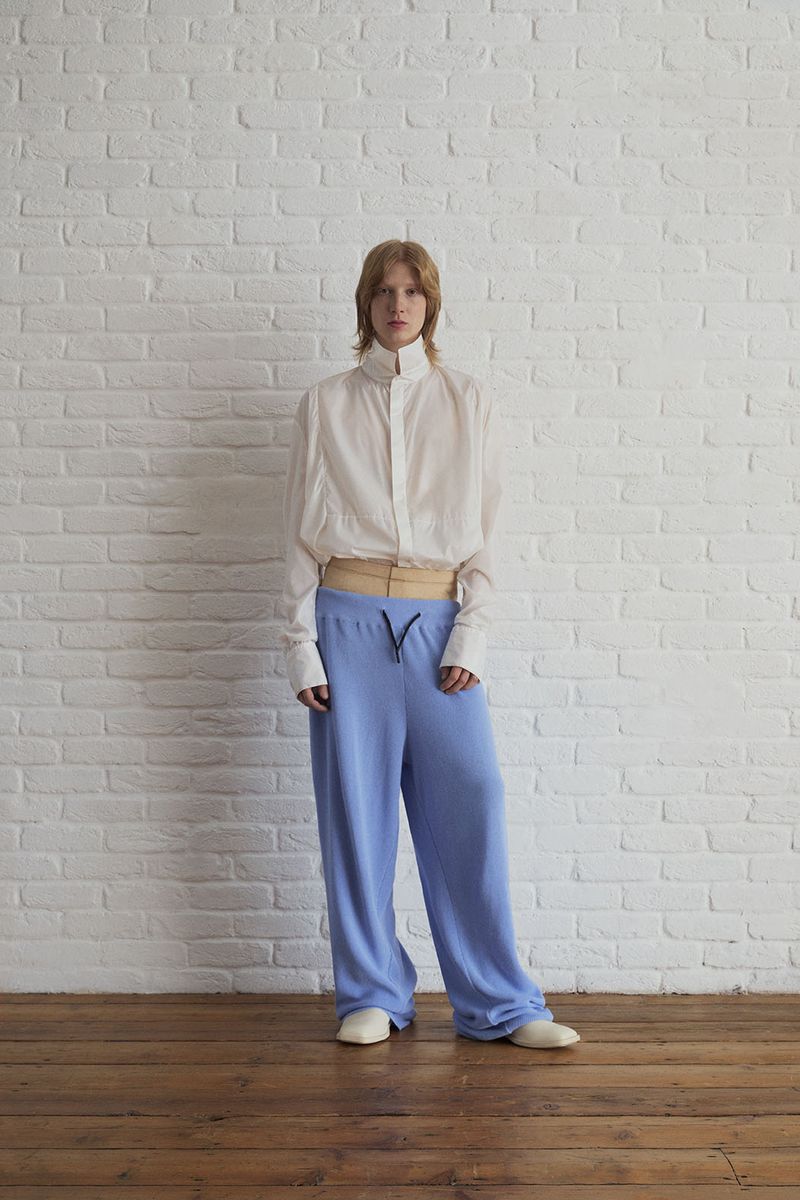 Full-body studio shot of a red-haired person in a white button-up shirt and blue sweatpants posing against a white brick wall.