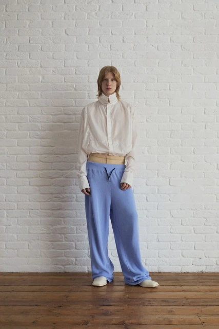 Full-body studio shot of a red-haired person in a white button-up shirt and blue sweatpants posing against a white brick wall.