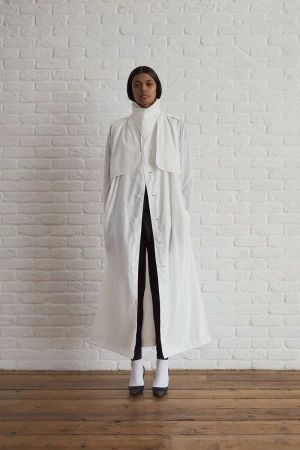 Elegant Black woman in long white coat and white boots poses against white brick wall in studio setting for a fashion editorial.