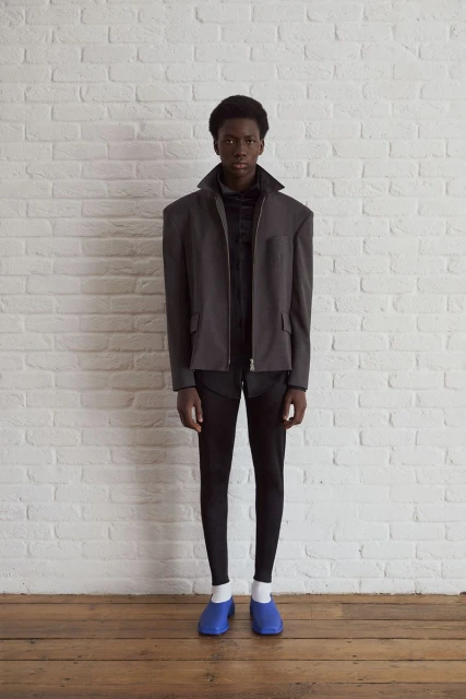 Fashion shot of an adult Black male in a grey blazer, black pants, and blue clogs standing indoors against a white brick wall backdrop.