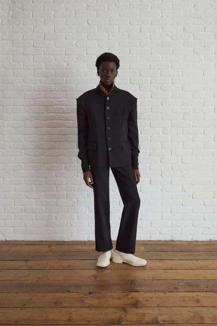 Adult male fashion model in a black suit and cream-colored shoes poses in a studio against a white brick wall and wooden floor.