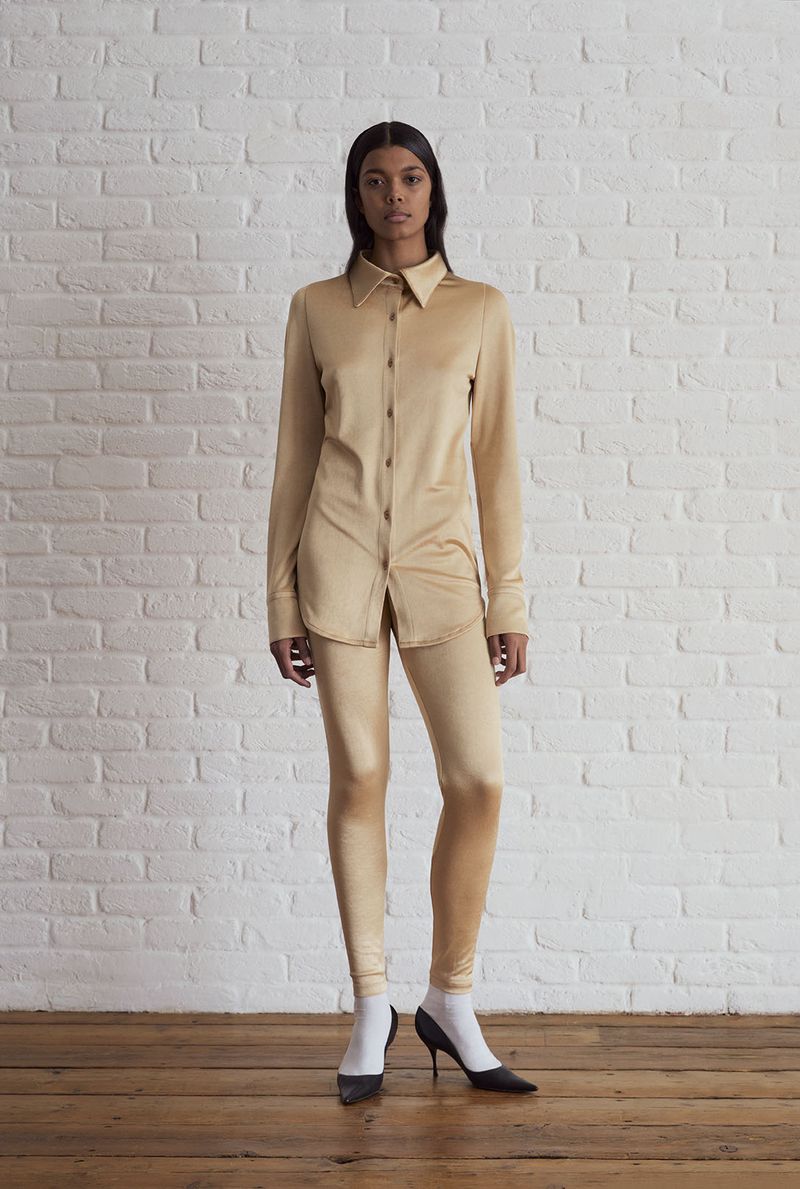 Woman in matching tan blouse and leggings stands against a white brick wall, wearing white socks and black pumps on a wood floor.