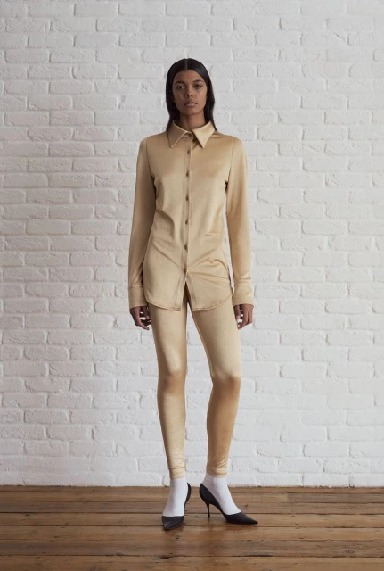 Woman in matching tan blouse and leggings stands against a white brick wall, wearing white socks and black pumps on a wood floor.