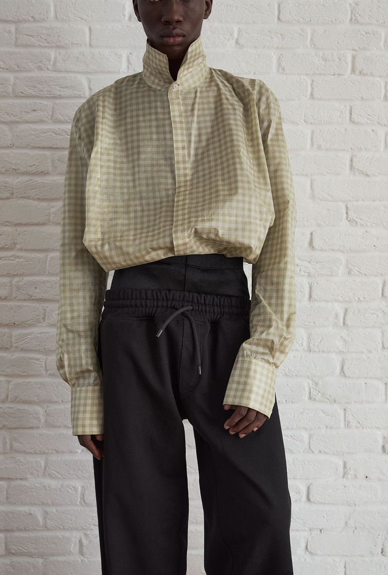 A slender Black person poses in a studio wearing a tan and white checked, long-sleeved top and black pants in a medium shot.
