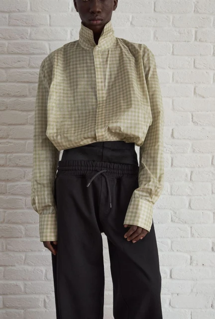 A slender Black person poses in a studio wearing a tan and white checked, long-sleeved top and black pants in a medium shot.