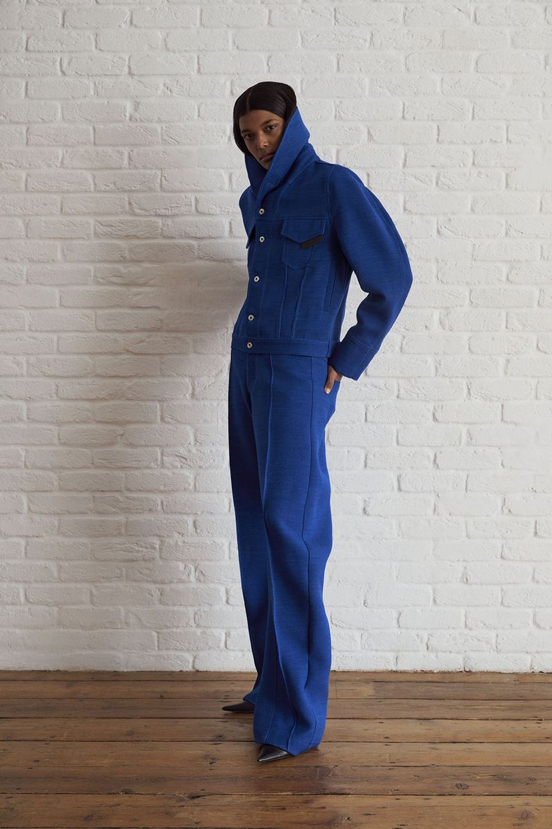 Woman poses in a bright blue jacket and matching pants in front of a white brick wall and a wooden floor. Full-length fashion shot.