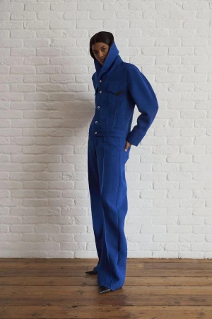 Woman poses in a bright blue jacket and matching pants in front of a white brick wall and a wooden floor. Full-length fashion shot.