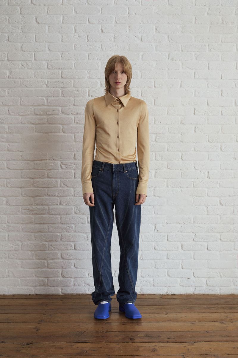 Full-length studio shot of a young adult man in tan shirt and blue jeans posing against a white brick wall with wood floor.