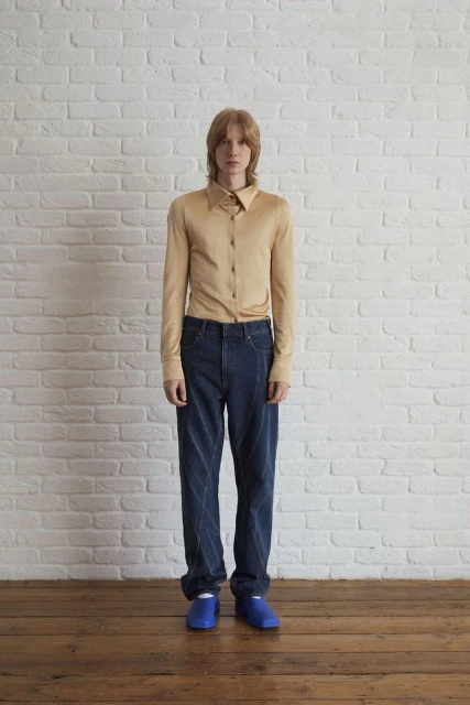 Full-length studio shot of a young adult man in tan shirt and blue jeans posing against a white brick wall with wood floor.
