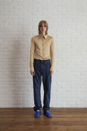 Full-length studio shot of a young adult man in tan shirt and blue jeans posing against a white brick wall with wood floor.