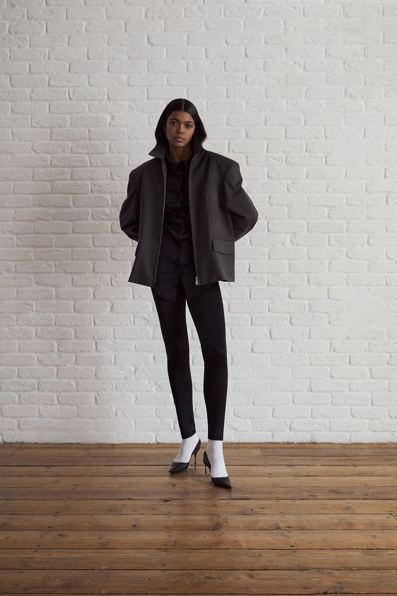 Model standing against a white painted brick wall wearing a structured dark jacket, black pants, white socks, and heels.