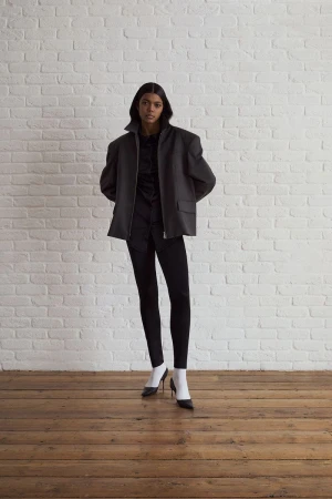 Model standing against a white painted brick wall wearing a structured dark jacket, black pants, white socks, and heels.