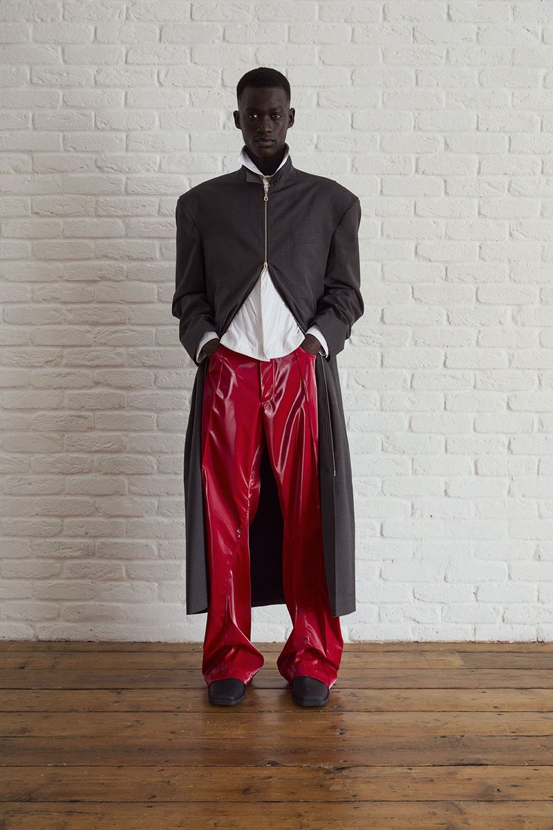 Full-length portrait of a Black male fashion model in an open grey coat, white shirt, red vinyl pants, against a white brick wall and wood floor.