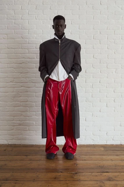 Full-length portrait of a Black male fashion model in an open grey coat, white shirt, red vinyl pants, against a white brick wall and wood floor.