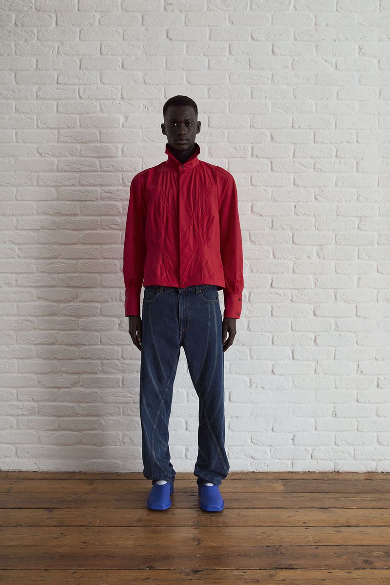 Dark-skinned adult male in a red collared shirt, blue jeans, and blue sneakers stands against a white brick wall on a wood floor.