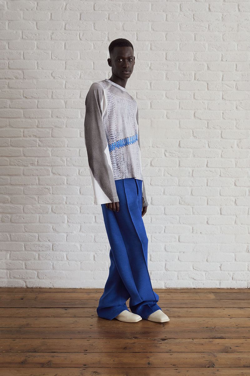 Fashion shot of a Black male model in a long-sleeved gray and white top and bright blue trousers, posing in front of a brick wall.