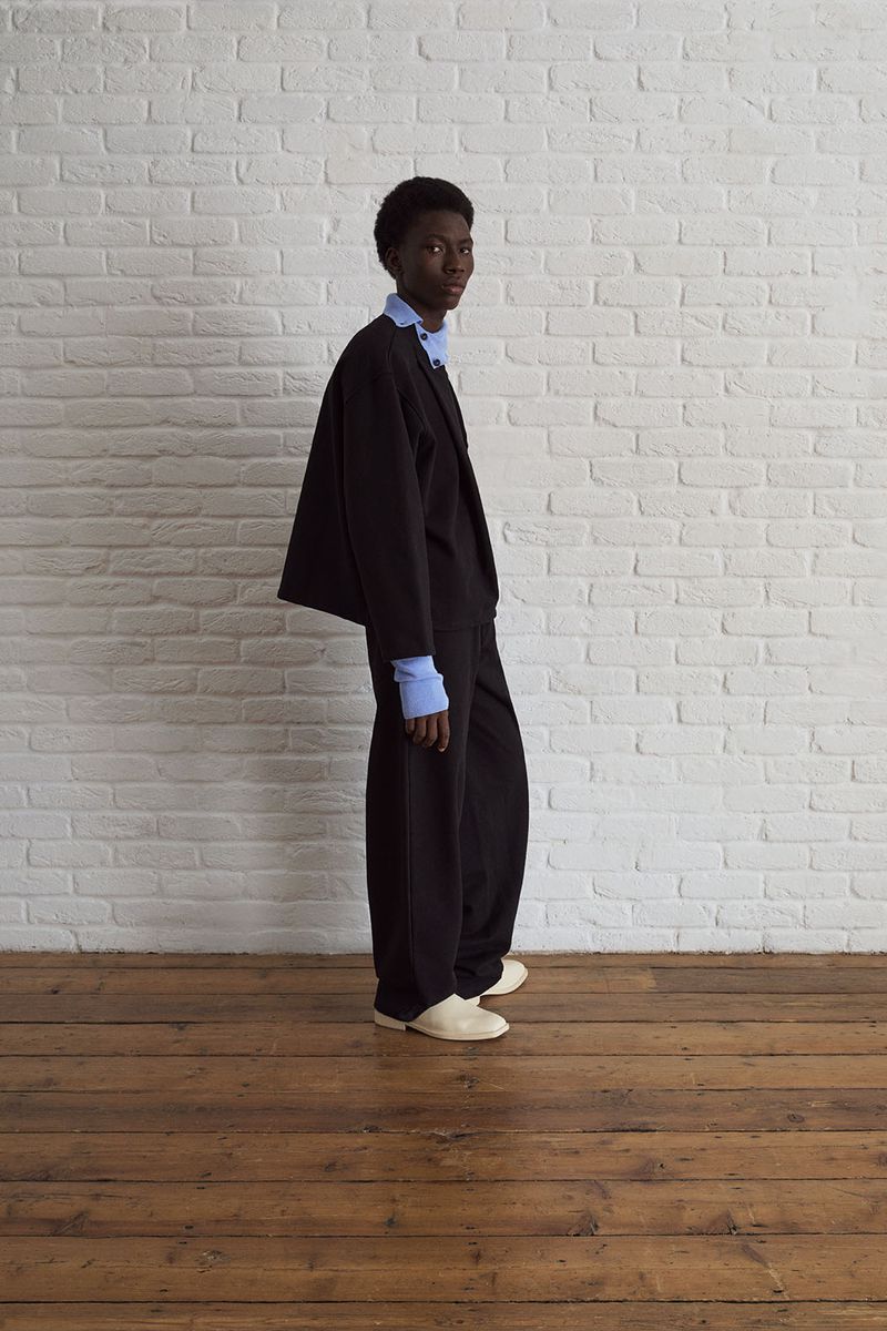 Dark-skinned adult male fashion model in dark suit and light-blue shirt against white-brick wall on brown wood floor in studio. 