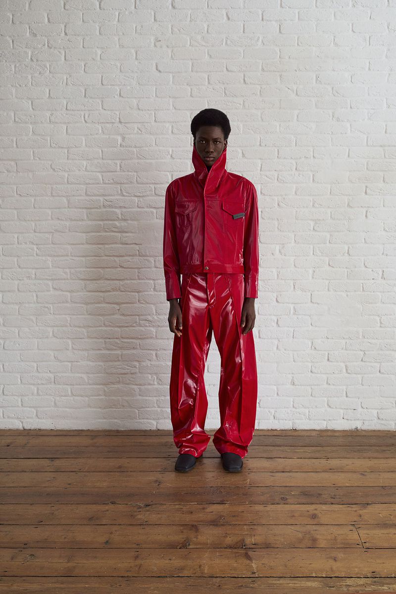 A young adult stands against a white brick wall wearing a bright red, shiny, futuristic-style two-piece outfit and dark shoes.