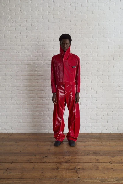 A young adult stands against a white brick wall wearing a bright red, shiny, futuristic-style two-piece outfit and dark shoes.
