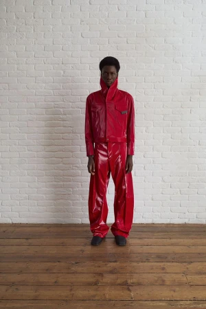 A young adult stands against a white brick wall wearing a bright red, shiny, futuristic-style two-piece outfit and dark shoes.