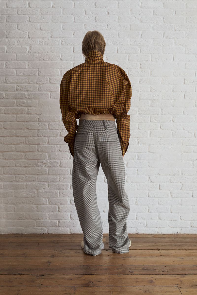 Person stands with their back to the camera in a plaid shirt and gray pants against a white brick wall and wooden floor.