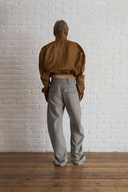 Person stands with their back to the camera in a plaid shirt and gray pants against a white brick wall and wooden floor.