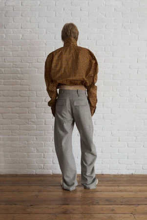 Person stands with their back to the camera in a plaid shirt and gray pants against a white brick wall and wooden floor.