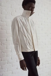 Black male model wearing a sculptural high-neck off-white pleated jacket and black trousers against a white painted brick wall.