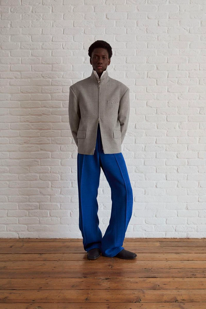 Black man standing against a white brick wall wearing a structured grey jacket and blue trousers.