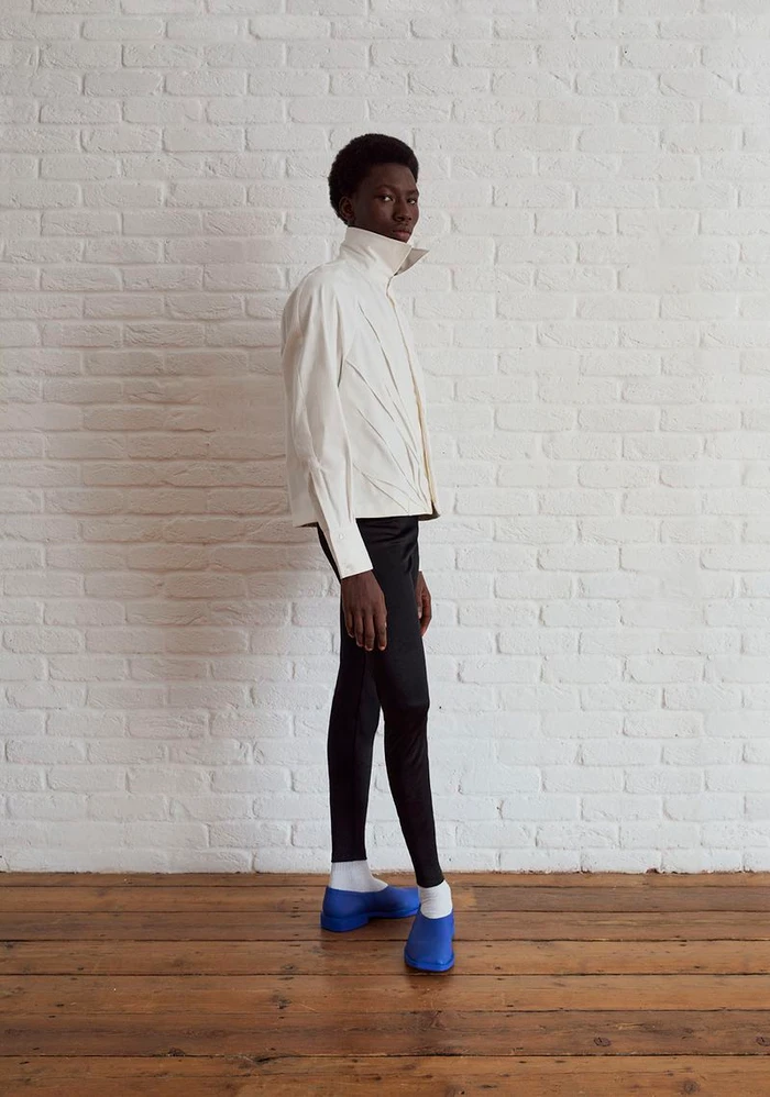 Model standing in profile wearing a cream high-collar jacket, black slim trousers, and bright blue shoes against a white brick wall.