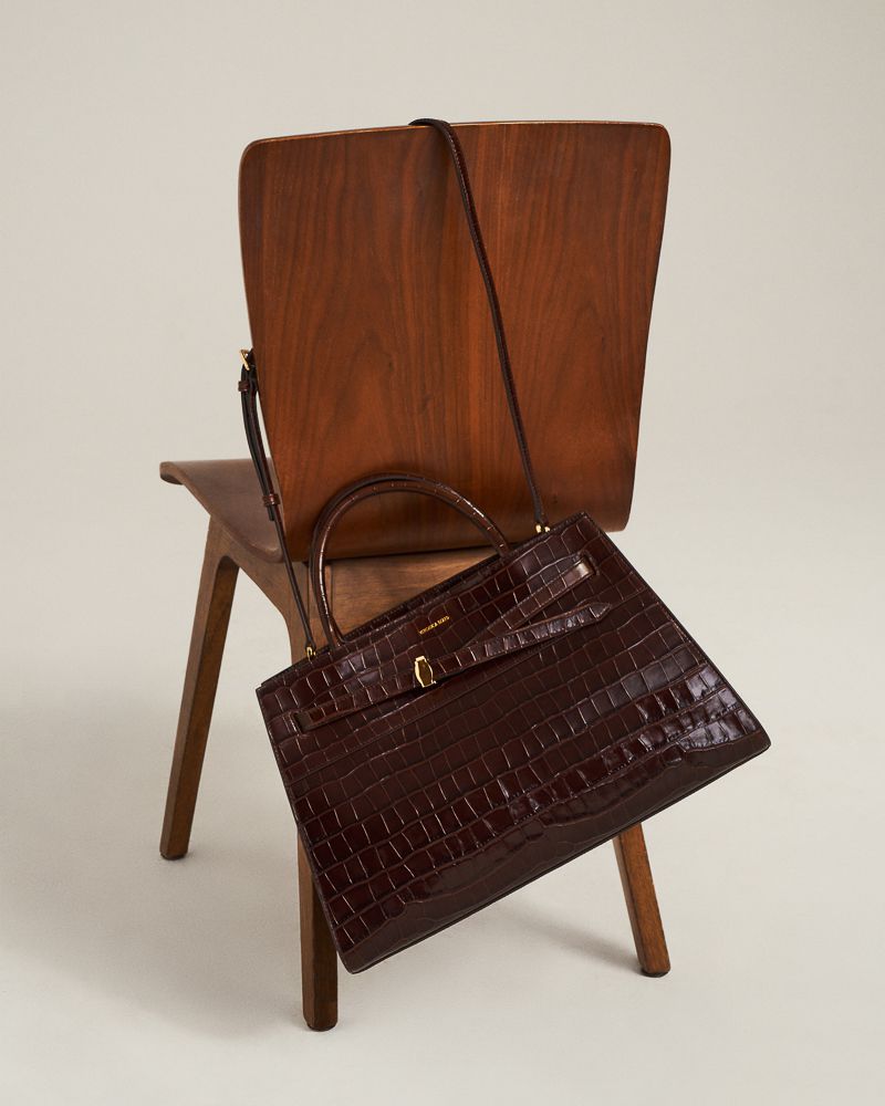 Brown faux crocodile handbag with a thin shoulder strap, displayed on a vintage wooden chair, against a cream-colored backdrop.