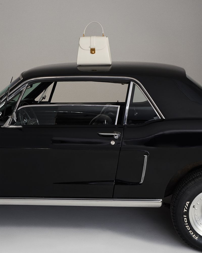 A white leather top-handle handbag sits on the glossy black roof of a vintage Ford Mustang coupe in a minimalist studio setting.