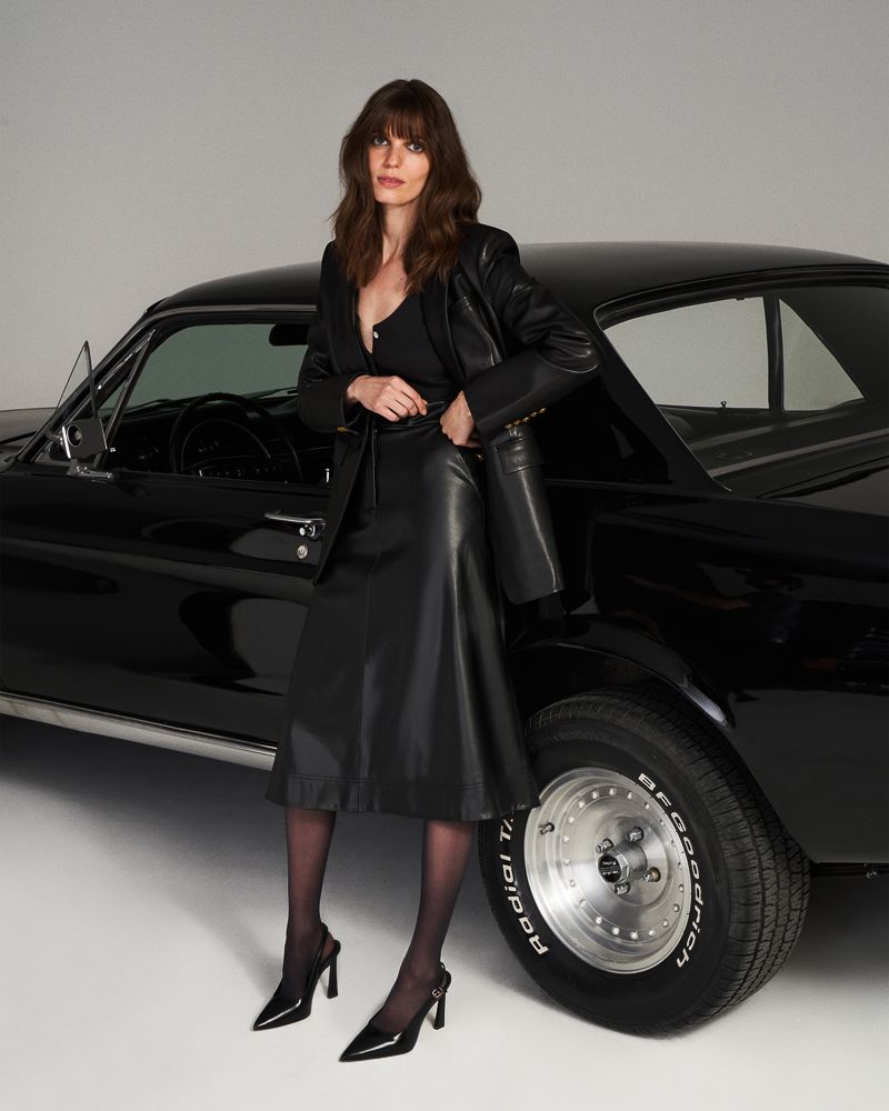 A brunette woman poses next to a black classic car wearing a black leather blazer and skirt, black hosiery, and black high heels.