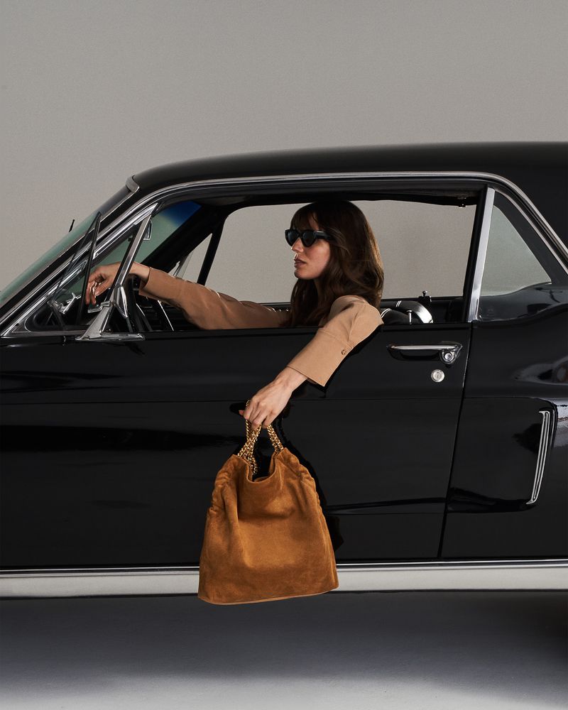 A woman in a black car holds a tan suede handbag with gold chain. She wears sunglasses and a tan blazer, with a neutral backdrop.