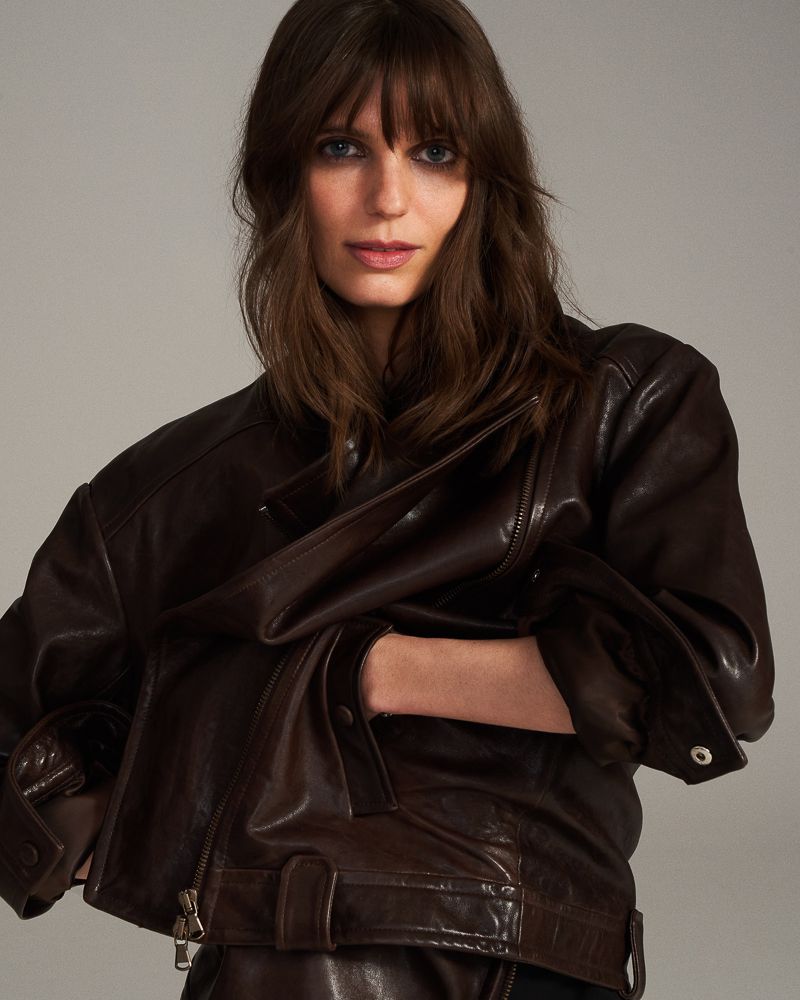 Portrait of a woman with brown hair and bangs wearing a dark brown leather biker jacket in a studio setting with a neutral background.