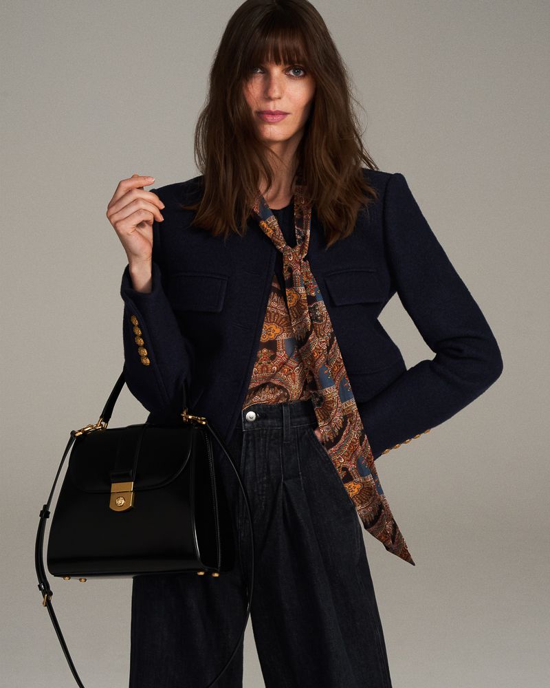 Brunette woman in a navy blazer, patterned top, and wide-leg jeans holds a black leather handbag in a studio fashion shot.