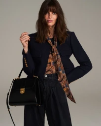 Model in dark blazer and paisley scarf holding a black handbag against a grey backdrop for Veronica Beard Fall 24.